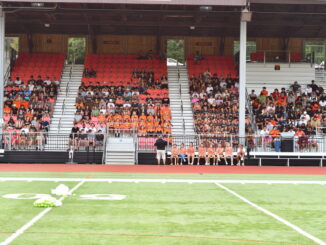 Students and coaches sitting in the bleachers across the football field.