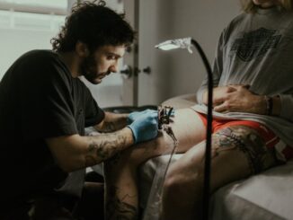 A white man wearing latex gloves tattoos a person's upper thigh.