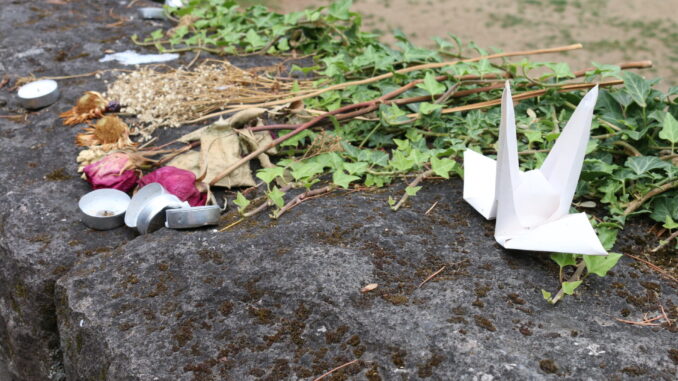 Flowers, candles, and a paper crane lay on top of a brick wall.