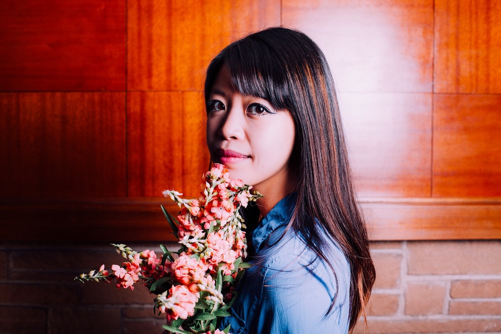 Image of Jane Wong staring at camera, holding a bouquet of flowers. brick background