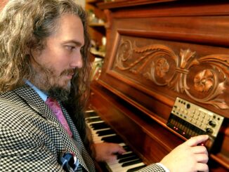 Image of Paul Evansmith in checkered suit sitting at a piano with one hand on the keys and another on a synthesizer propped up on the piano