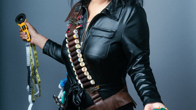 Image of Kristina Wong wearing leather and posing holding a pin cushion that looks like a bomb, measuring tape, needle and thread and other sewing supplies likened to weapons