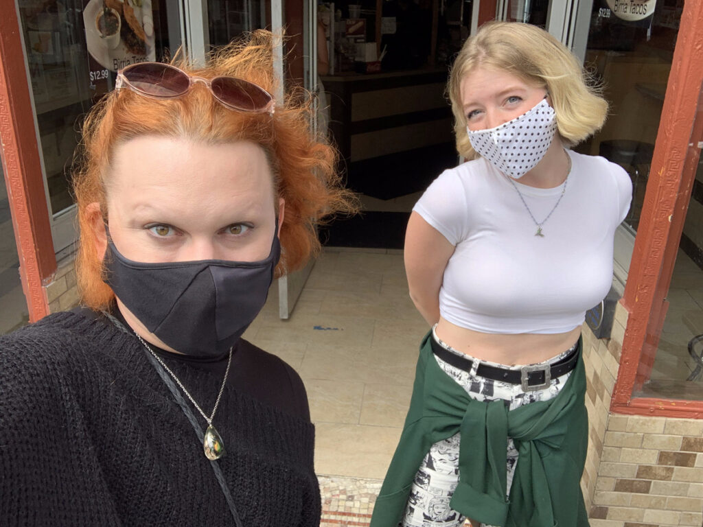 Paige stands smililng behind Jinkx Monsoon who is taking a selfie of them. both are wearing cloth masks.