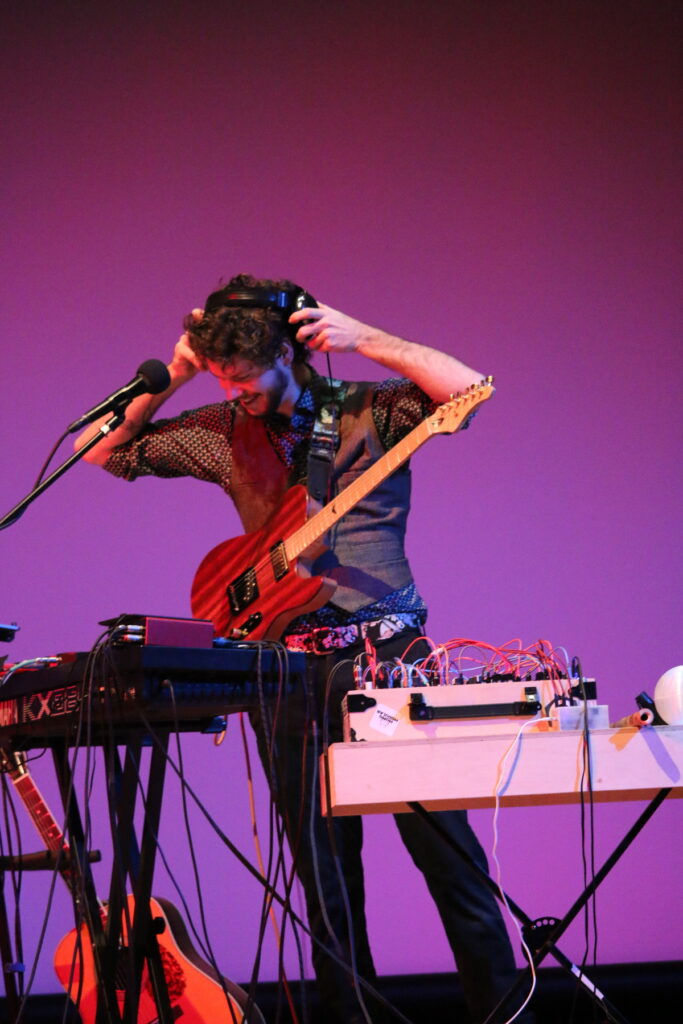 Max Krien adjusting his headphones onstage in front of a sound station, while wearing an electric guitar