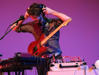 Max Krien adjusting his headphones onstage in front of a sound station, while wearing an electric guitar