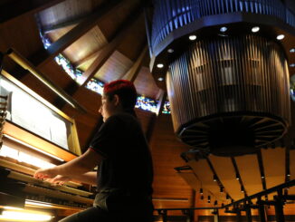 Photo of Grace playing organ in foreground, with circular suspended portion of organ seen behind him