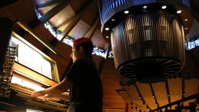 Photo of Grace playing organ in foreground, with circular suspended portion of organ seen behind him